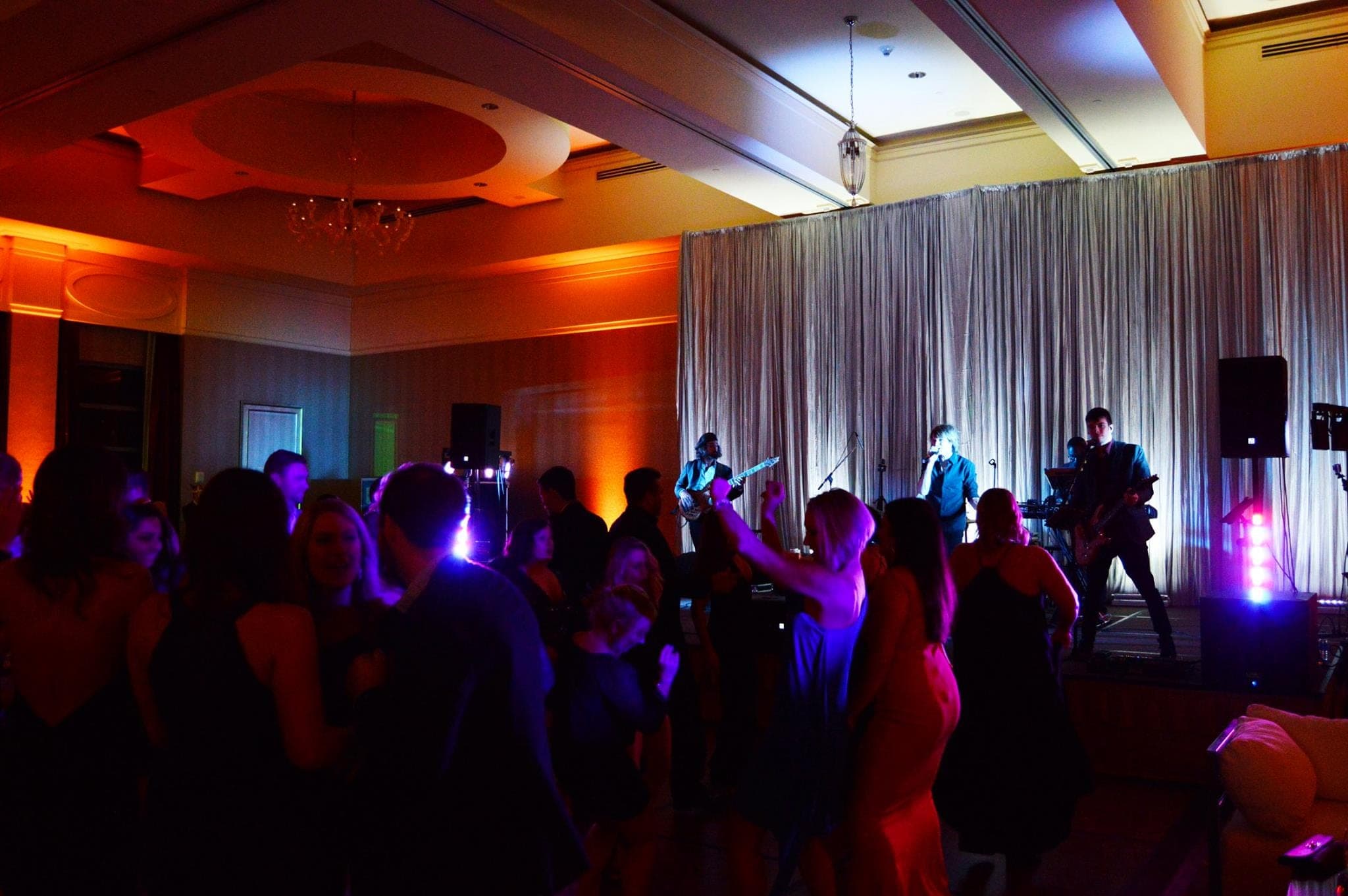 A ballroom dance floor full of guests under purple light, with the band on stage behind them